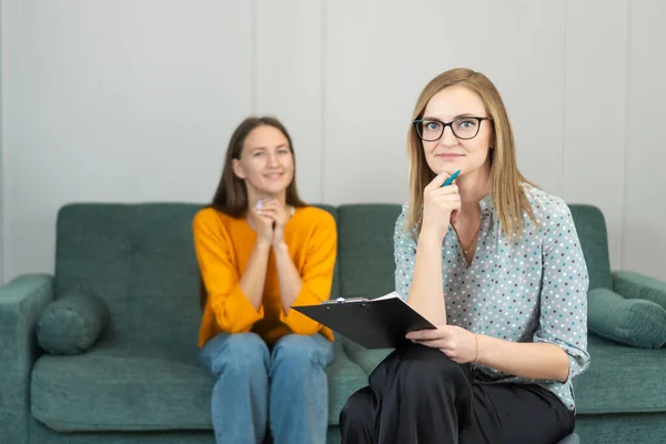 stock image Smiling female psychologist with glasses, leaning her hand on her chin, looks into the camera, consulting a young woman in the office interior. Therapy, psychological support, professional help