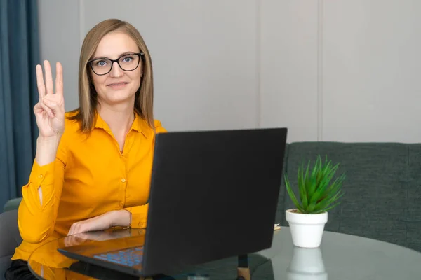 Business woman with a smile in an orange shirt and glasses is sitting at a desk in the office with a laptop, pointing fingers number three.
