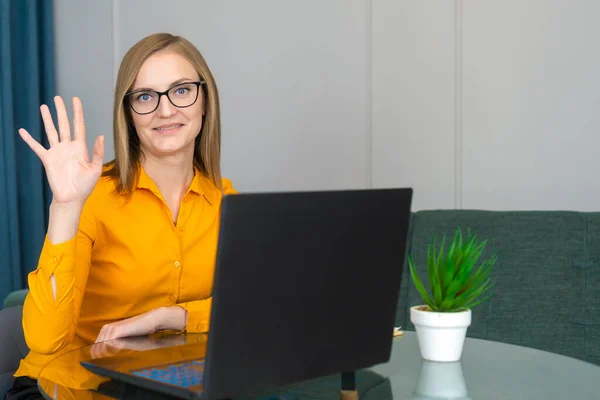 Business woman with a smile in an orange shirt and glasses is sitting at a desk in the office with a laptop, pointing fingers number five.