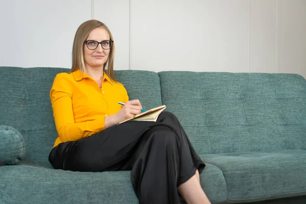 Woman in an orange shirt, black trousers and glasses is sitting on the couch taking notes in a notebook. Business woman with glasses looks into the camera. Business, journalist, psychologist, coach