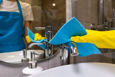 Sarı lastik eldivenli bir kadının eli modern bir banyoda musluğu mavi bir temizleme beziyle silerken. Profesyonel temizlik kavramı