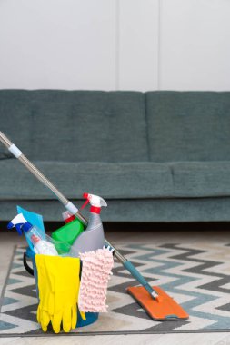 Mavi bir kovanın içinde temizlik ürünleri, kimyasallarla dolu şişeler, lastik eldivenler, bir bez parçası, kanepenin önünde duran bir bez parçası, kopyalama alanı. Temizlik servisi. Dikey fotoğraf