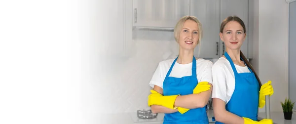 两个身穿蓝色制服 面带微笑的年轻女子站在厨房里 看着摄像机 一个手臂交叉在胸前 另一个拿着拖把 清洁的概念 — 图库照片