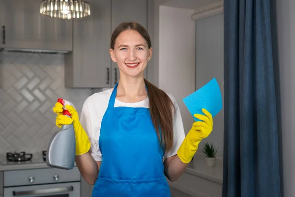 Smiling young woman in a blue uniform apron,yellow rubber gloves holds a spray and a microfiber cleaning cloth in her hands.Hiring a qualified home care agency, a quick and simple housekeeping concept