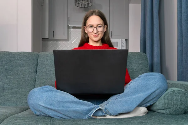 안경을 매력적 쇼파에서 노트북을 만들고 있습니다 스웨터를 아름다운 미소짓는 소녀는 — 스톡 사진