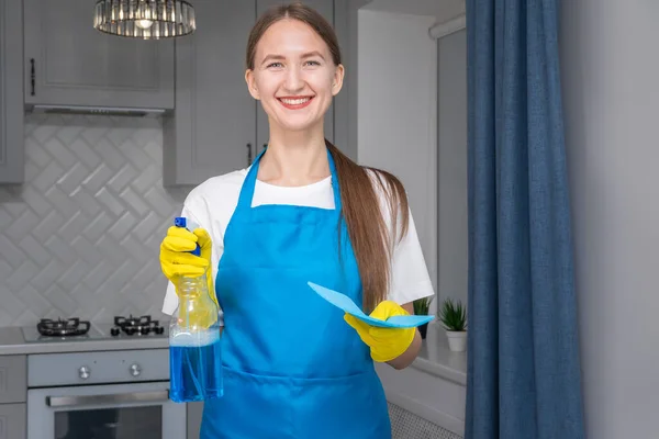 Young smiling woman in a uniform apron holds a bottle of detergent and a napkin. Hiring a qualified home care agency, a quick and simple housekeeping concept. Cleaning company. House cleaning.