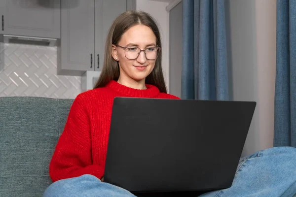 안경을 매력적 쇼파에서 노트북을 만들고 있습니다 스웨터를 아름다운 미소짓는 소녀는 — 스톡 사진