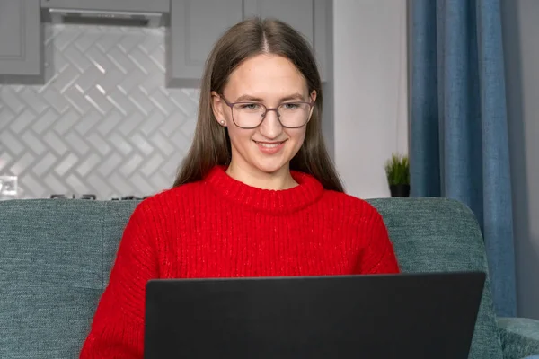 穿着红色毛衣 牛仔裤 眼镜的快乐微笑的年轻女人 带着笔记本电脑坐在沙发上 漂亮的女孩在网上工作 购物或交流 — 图库照片