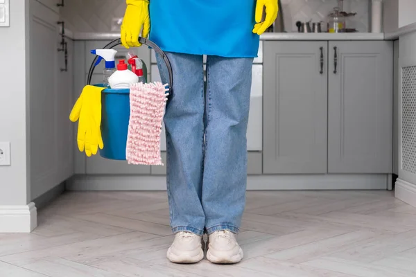 stock image Uniformed cleaner holds in her hands a blue bucket filled with chemicals, cleaning products, a rag standing against the background of the kitchen. Cleaning company concept, house cleaning