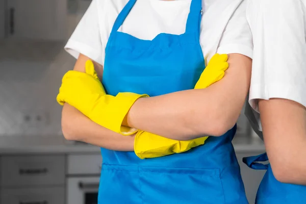Cleaning team of two people with their arms crossed on their chests against the background of a blurry kitchen. Professional female workers in aprons and yellow rubber gloves cleaning company.