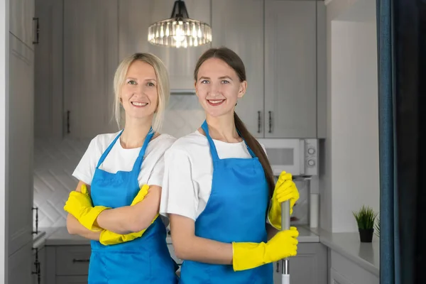 Two young beautiful smiling women in aprons are standing shoulder to shoulder with their arms crossed on their chests looking at the camera. Conceptual advertising of a cleaning company