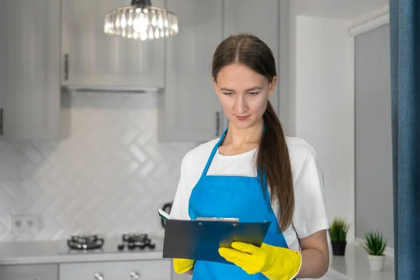 Young beautiful professional cleaning woman in an apron from a cleaning company checks the work done using a checklist on a paper planter or accepts an order to perform house cleaning work