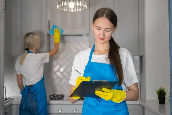 Young woman from a professional cleaning company in uniform notes and writes down a list of the work done on cleaning the room in a tablet. Stove is being laid in the background.