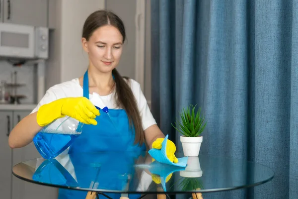 Beautiful young woman in an apron and yellow rubber gloves sprays and wipes a glass table with a microfiber cloth. Concept of housework. Cleaning company