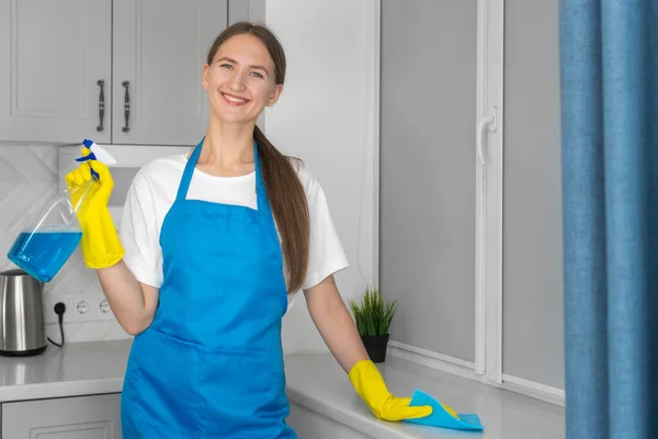 Smiling young beautiful woman in a blue apron, yellow rubber gloves is standing near the window with detergent and a microfiber cloth. Cleaning concept
