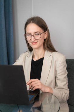 Gözlüklü zeki, güzel, çekici ve çekici genç bir kadın bir dizüstü bilgisayarla online toplantı düzenliyor. Hafif ceketli bir iş kadını laptopta masada oturuyor. Dikey fotoğraf
