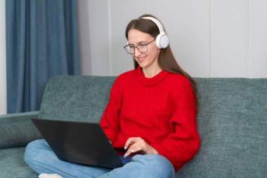 Kırmızı kazaklı, beyaz kulaklıklı, mutlu, genç bir kadın elinde dizüstü bilgisayarla kanepede oturuyor, gülümsüyor. Çevrimiçi iletişim, eğitim, uzaktan çalışma kavramı
