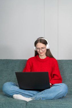 Kırmızı kazaklı, kot pantolonlu, beyaz kulaklıklı genç bir kadın evdeki kanepede internette geziniyor. Ev ofisinde çalışan bir kız. Bir dizüstü bilgisayar ve internet kullanan serbest çalışan. Oturma odasındaki işyerinde.