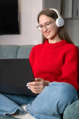 Kırmızı kazaklı, kot pantolonlu, beyaz kulaklıklı genç bir kadın evdeki kanepede internette geziniyor. Ev ofisinde çalışan bir kız. Bir dizüstü bilgisayar ve internet kullanan serbest çalışan. Oturma odasındaki işyerinde.