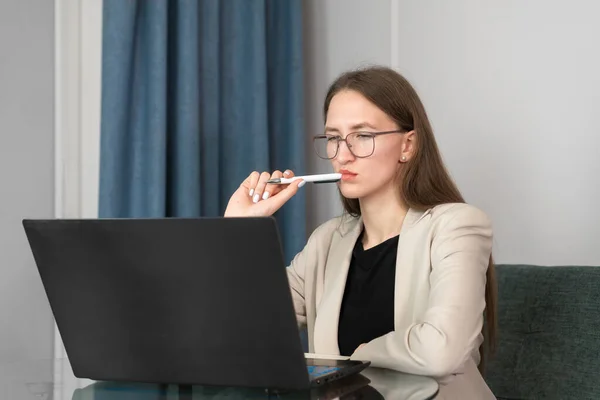 眼镜是一位穿着夹克 充满热情的女商人的年轻雇员 她坐在玻璃桌旁 嘴边拿着一支笔 看着笔记本电脑 职业理念的成就 自由职业 — 图库照片
