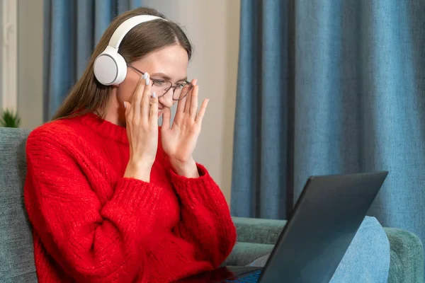 可爱的年轻的棕色头发的女人穿着一件红色毛衣 白色耳机调整她的眼镜 当她坐在客厅的沙发上看笔记本电脑时 远程工作 自由职业者 — 图库照片
