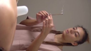 Kadın masör, yanan çubuğun arka planında hasta masajı yapıyor dumanlı, aromaterapili. Spa odası, masaj masası, havlular, kozmetik ürünleri, masaj yağları, tütsü çubukları. Dikey video