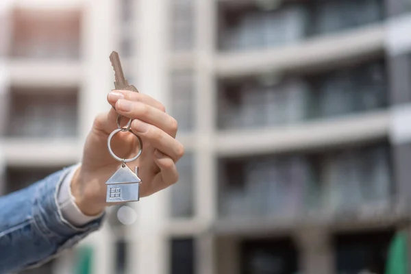 stock image Close-up of a woman's hand with a key and a keychain of a house against the background of a blurred modern residential complex. Concept of buying real estate, apartments