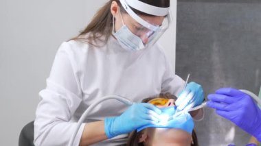 Dişçi kadın klinikte genç bayan hastaların dişlerini tedavi ediyor. Diş hekimi diş matkabı ve diş cilası kullanırken yüz maskesi takar. Modern dişçi muayenehanesinde diş prosedürleri.