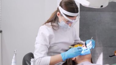 Dişçi kadın klinikte genç bayan hastaların dişlerini tedavi ediyor. Diş hekimi diş matkabı ve diş cilası kullanırken yüz maskesi takar. Modern dişçi muayenehanesinde dişçi prosedürleri. Lateks peçete. lastik.