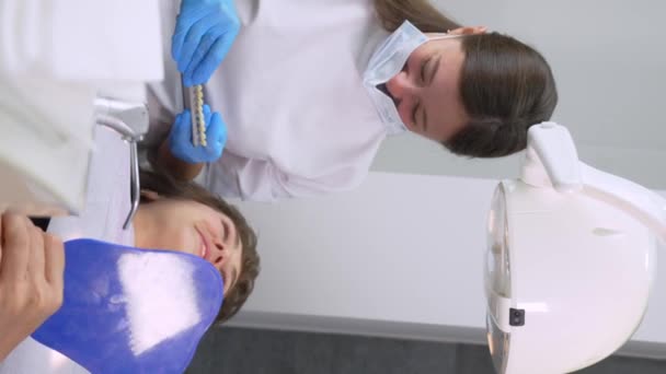 女の子の患者と入れ歯の歯の日陰の選択 歯医者は特別なスケールから歯の色を選択します 歯科手術の椅子に座っている若い女性 プロソドトニクス 垂直ビデオ — ストック動画