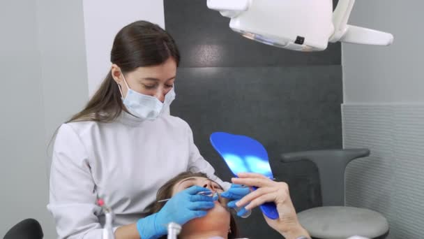 幸せな若いヨーロッパの女性は 歯科医院で自分自身を鏡で見て予約中に歯科医と甘い通信し 話をします 現代医学 女性は笑顔で問題について医者に話す — ストック動画