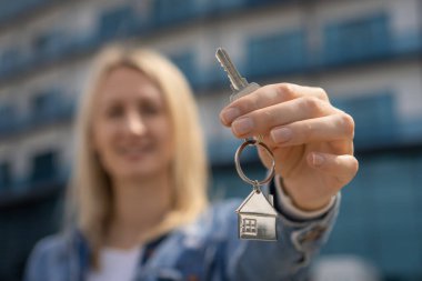 Sarışın genç bir kadın, yeni bir evin arka planında caddede duran, uzanmış elinde bir anahtar tutuyor. Gayrimenkul konsepti, aracı, alıcı