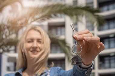 Genç bir kadın, modern yeni bir evin ve palmiye ağaçlarının arka planına karşı bir anahtarla elini uzattı. Emlakçılık. Mutlu komisyoncu. Emlak satın almak
