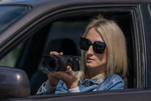 stock image Young blonde woman in sunglasses takes pictures on a professional camera while sitting in a car. Concept of journalism, detective, papparation
