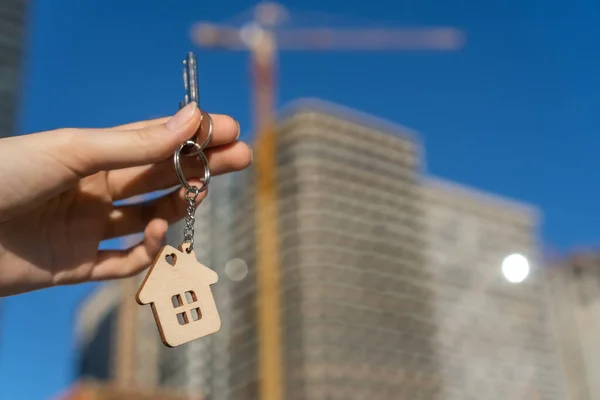Woman\'s hand holds key and keychain in form of house against background of house under construction. concept of own housing. Buying apartment from real estate agency mortgage. Housewarming settlement