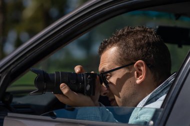 Man in sunglasses with camera sits inside car and takes pictures with professional camera, private detective or paparazzi spy. Journalist seeks sensation and follows celebrities. clipart