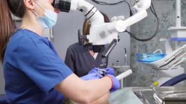 Diş mikroskobuyla çalışan iki kadın dişçi. Doktor ve asistanı dişçi kliniğinde. Doktor mikroskoba bakıyor ve elinde diş ve ayna tutuyor. Modern diş kliniği. Diş hekimliği.