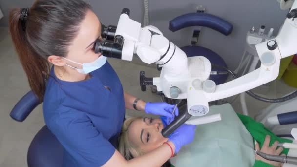 歯科顕微鏡を用いた女性歯科医歯クリニック事務所 顕微鏡を使った経験豊富な歯科医による治療を受けています 歯医者さんの椅子の上の女性のイメージ — ストック動画