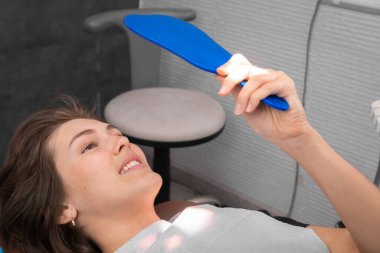 Genç, gülümseyen bir kadın dişçinin ofisindeki sandalyede aynada dişlerine bakar. Mutlu, dişçiye gittikten sonra hastanın sonucundan memnun.