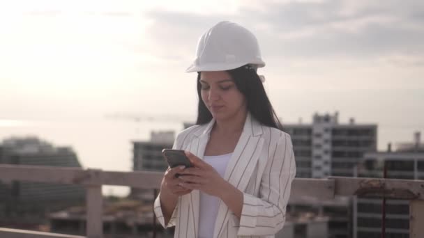 ヘルメットの建設現場の女性建築家は携帯電話を保持しています 青い空と建設現場を背景に建築家として活動 美しい女性の顔 — ストック動画