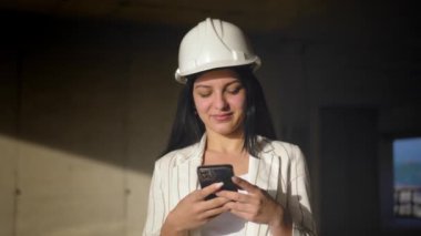 İnşaat alanındaki miğferli kadın mimar cep telefonu tutuyor. Müteahhidin mimarı mavi gökyüzü ve inşaat sahasının zeminine karşı çalışıyor. Güzel Dişi Yüz. Dikey video.