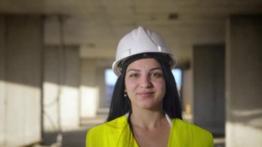 İnşaat alanında kask ve yeşil yelek giyen orta yaşlı kadın mimar çalışırken gülümsüyor ve kameraya doğru yürüyor. Başarılı ve gururlu müfettiş. Kadın Mühendis Portresi. Başı