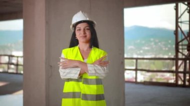 Miğfer ve yeşil yelek giyen orta yaşlı kadın mimar kollarını göğsünde kavuşturuyor. İnşaat alanında çalışırken gülümsüyor. Başarılı ve gururlu müfettiş. Kadın Mühendis Portresi. Başı
