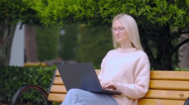 Çevrimiçi iş kadını. Genç bir kadın dizüstü bilgisayarla kentteki bankta oturuyor, yazı yazıyor ve güneşli bir günde çalışıyor. Eğitim kavramı. Serbest çalışan biri. Sınav hazırlığı için öğrenci bilgisayarı. Gözlüklü.