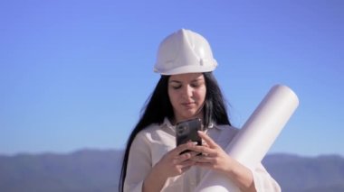 İnşaat alanındaki mimar kadının elinde cep telefonu ve katlanmış kağıt var. Geliştiricinin mimarı mavi gökyüzünün arka planına karşı çalışıyor. Güzel kadın yüz şirketi..