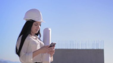 İnşaat alanındaki mimar kadının elinde cep telefonu ve katlanmış kağıt var. Geliştiricinin mimarı mavi gökyüzünün arka planına karşı çalışıyor. Güzel kadın yüz şirketi..