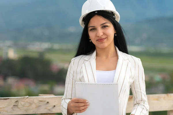 年轻美丽的 面带微笑的高加索女建筑师 头戴防护头盔 身穿夹克 站在建筑工地上 用的是纸片 — 图库照片