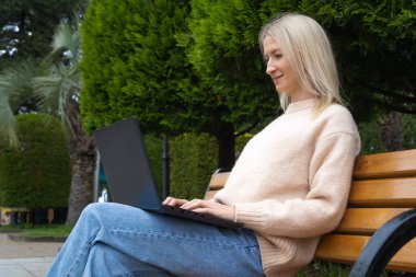 Parkta dizüstü bilgisayarla bankta oturan güzel bir sarışının yan görüntüsü..