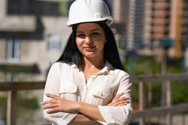 Güneşli bir günde inşaat sahasında kasketli genç ve güzel bir kadın mühendis elleri önünde katlanmış kameraya bakıyor..