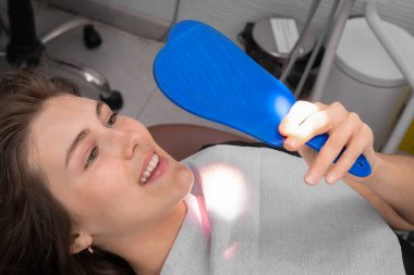 Genç, gülümseyen bir kadın dişçinin ofisindeki sandalyede aynada dişlerine bakar. Mutlu, dişçiye gittikten sonra hastanın sonucundan memnun.